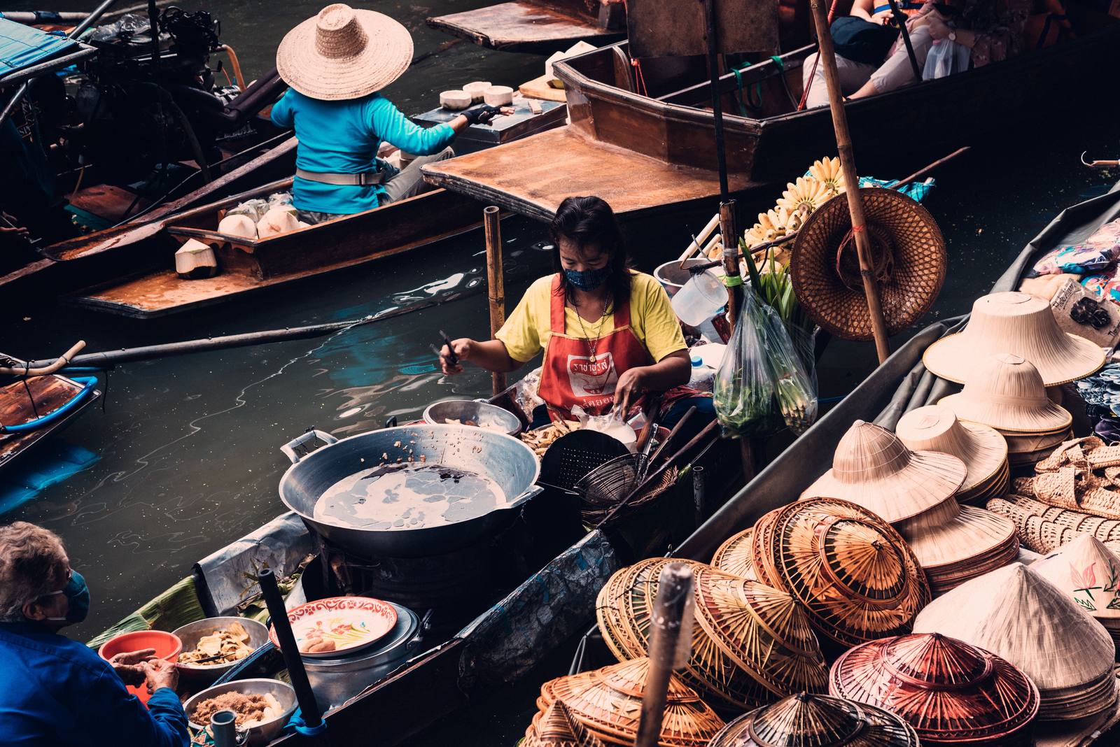 Vendedora preparando comida en su barca en un mercado flotante de Tailandia, rodeada de sombreros de bambú y fruta