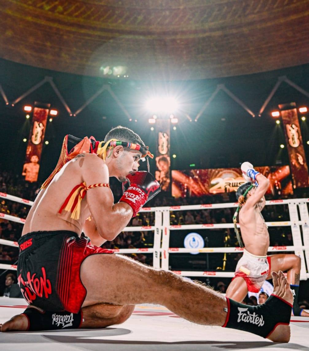 Combate de Muay Thai bajo los focos de un estadio, con los luchadores realizando el ritual Wai Kru Ram Muay